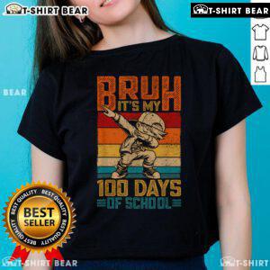Boy wearing 'Top Bruh It's My 100 Days of School' tee celebrates 100th day of school in a colorful classroom.